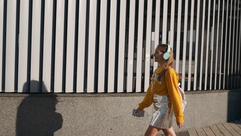 Portrait of young cute attractive young girl in urban background listening to music with headphones. Woman wearing yellow blouse and silver skirt.