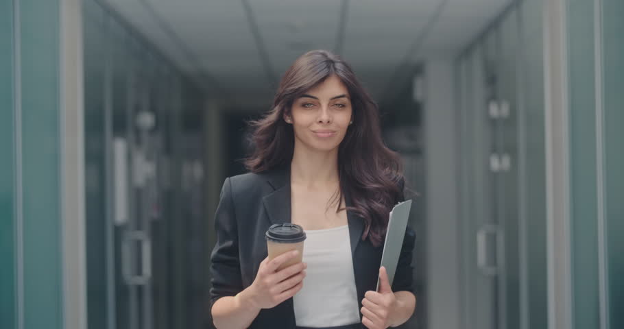 Portrait of young successful businesswoman walking in office hall with pad and cup of coffee in her hands, looking at camera and smiling 4k Royalty-Free Stock Footage #1013074283