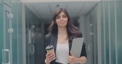 Portrait of young successful businesswoman walking in office hall with pad and cup of coffee in her hands, looking at camera and smiling 4k