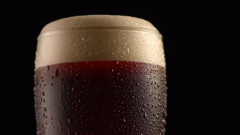 Dark Beer closeup. Pint of cold Craft beer isolated on matte black background, rotation 360 degrees. Glass of beer with water drops. 4K UHD video 3840x2160