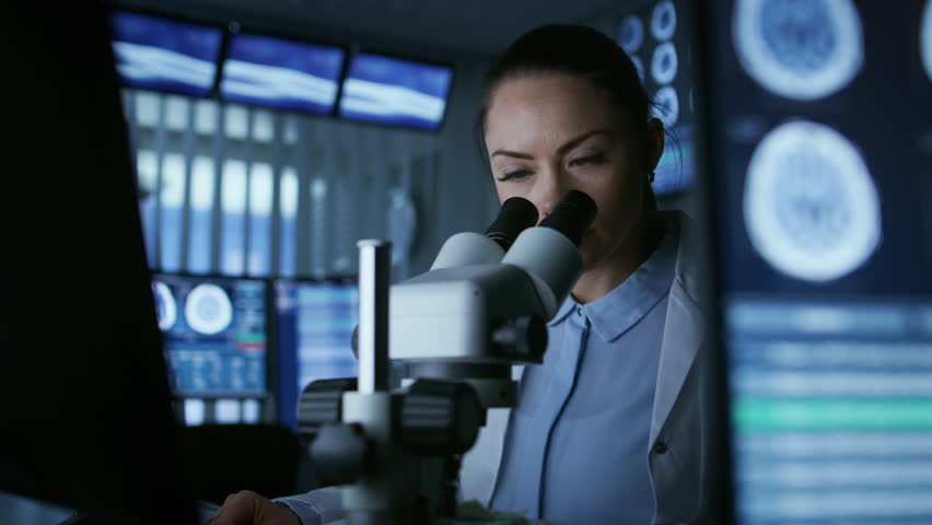 Female Medical Research Scientist Looking Through the Microscope Types Acquired Data in the Computer. Laboratory.  Shot on RED EPIC-W 8K Helium Cinema Camera.