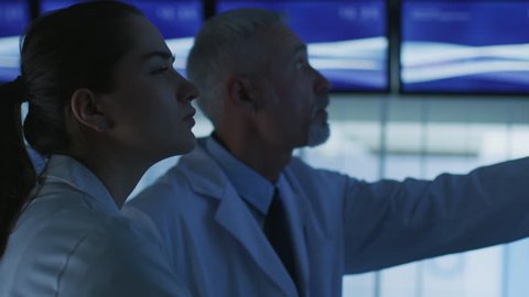 Close-up Shot of Two Medical Scientists  Neurologists, Talking and Pointing at TV Wall Monitor in the Modern Laboratory. Research Scientists Making New Discoveries in the fields of Neurophysiology.