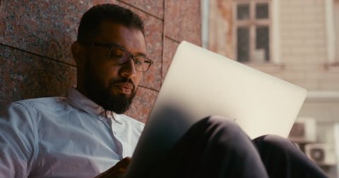 Close-up portrait of the handsome focused african businessman in eyeglasses typing on the laptop outdoor.