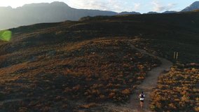 aerial drone footage behind a male trail runner running along a single track path through a mountain landscape at sunset