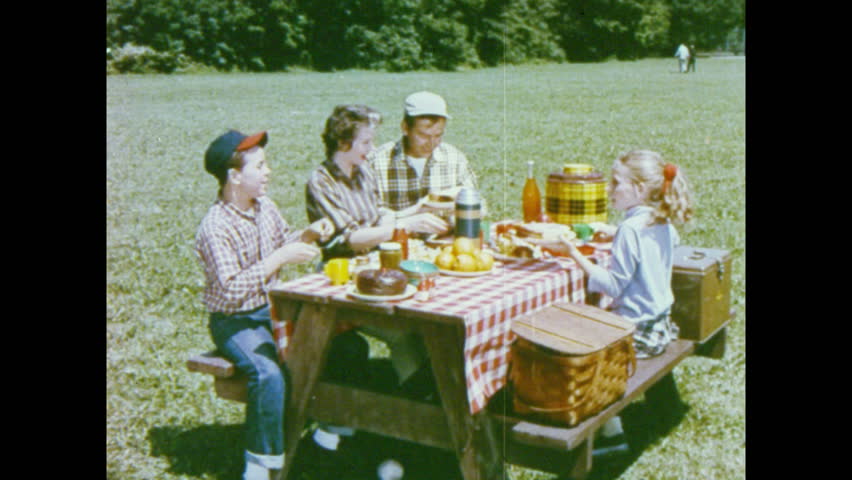 1950s: Family sits around picnic table, passes food around, makes sandwiches, talks. Boy eats sandwich, girl eats sandwich.