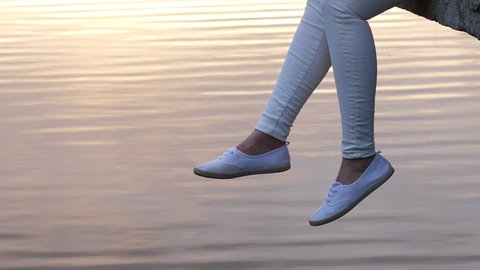 Woman legs in white shoes dangle over calm water surface, recreating concept