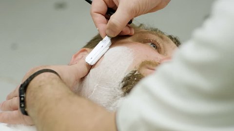 Shaving of beard. Barber cutting men's face hair with straight razor at barbershop. 4K