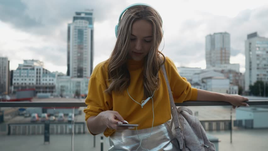 Portrait of young cute attractive young girl in urban city streets background listening to music with headphones. Woman wearing yellow blouse and silver skirt. | Shutterstock HD Video #1013167247