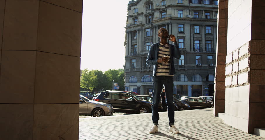 Slow motion of the African American smiled young man standing in the town on a sunny day and speaking on the phone with a coffee-to-go in hands. Inside.