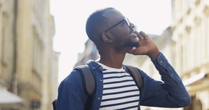 Close up of the African American young man in the jeans shirt and in glasses talking on the phone on the street on a sunny day. Outdoors.