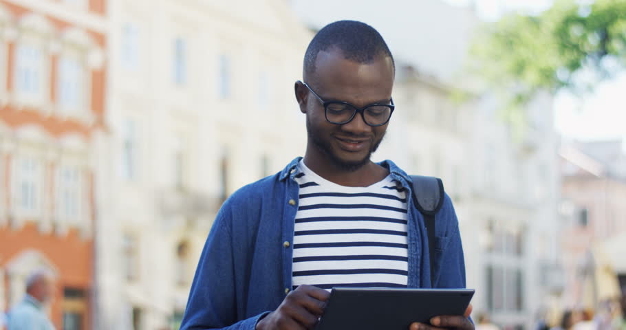 Portrait of the African American male tourist with a backpack standing on the street of a town, looking around and scrolling, taping or texting on the tablet. Outdoor.