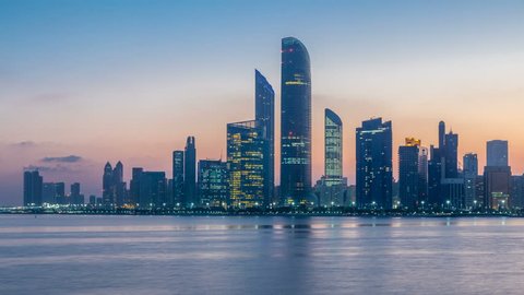 Abu Dhabi city skyline with skyscrapers before sunrise with water reflection night to day transition timelapse from the Breakwater near cultural village. Few clouds on morning sky