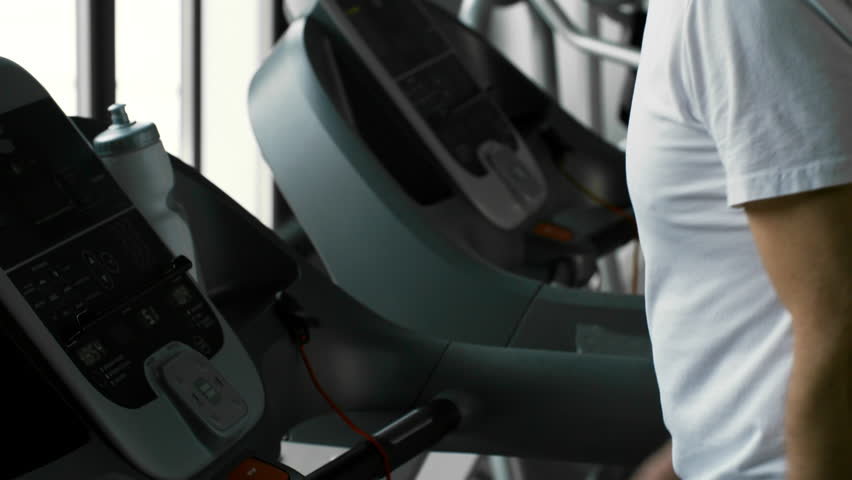 Medium shot of thirsty mature man taking bottle and drinking water while doing brisk walk on treadmill at the gym