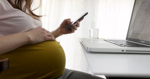 Crop side view of casual pregnant woman sitting at table and using smartphone
