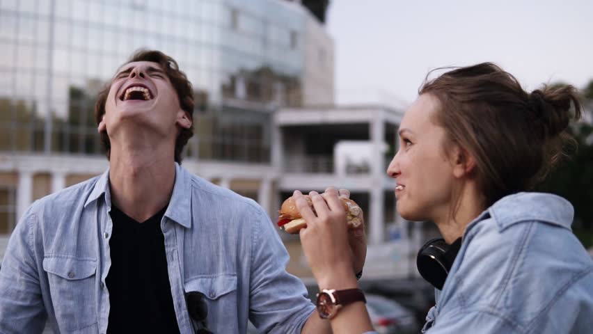 Happy, young couple are hanging out in the park. Lauging and smiling. Girl is eating burger. The young guy gently wipes the sauce off his girl's lips and licks a finger. Close up