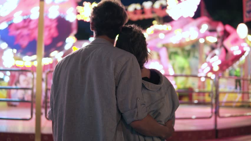 The loving couple is standing and looking at the luminous attraction at the amusement fair. Glare of light, night