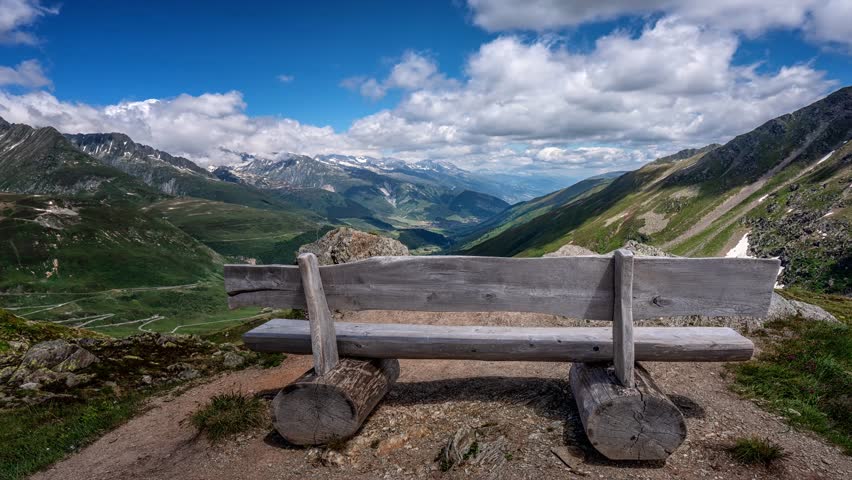 Timelapse of an empty wooden bench overlooking a valley in the Canton Graubünden Switzerland with clouds and blue skies and lush green grass on mountains