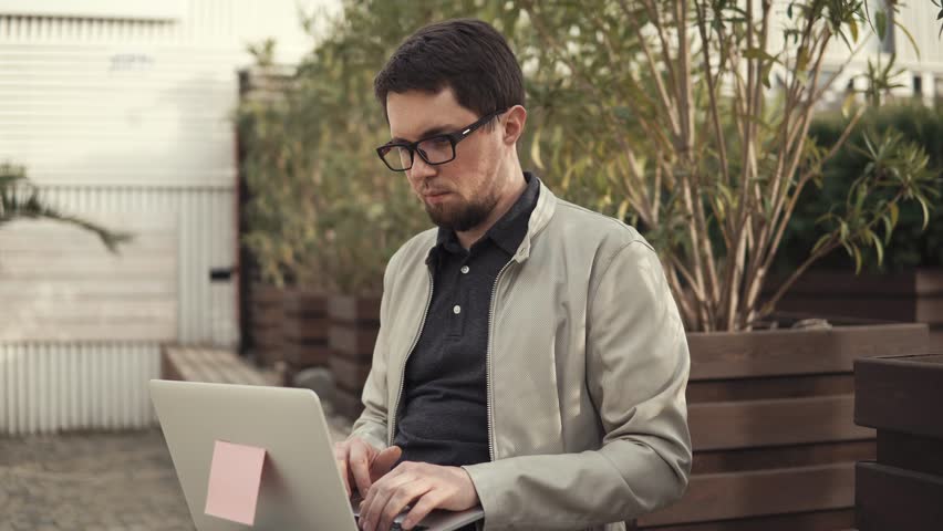 Young businessman with poor eyesight developing a startup project on a laptop, a gentleman holding a device on his lap sitting on a park bench | Shutterstock HD Video #1013189879