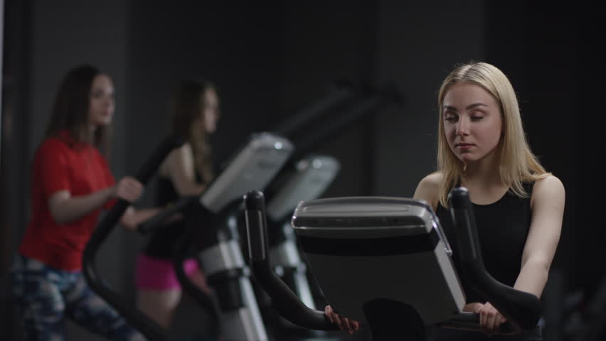 beautiful girl in blak sport wear vigorously works on exercise bike and coach instructs about right way of exercise in the new gym against the background of engaged girls
