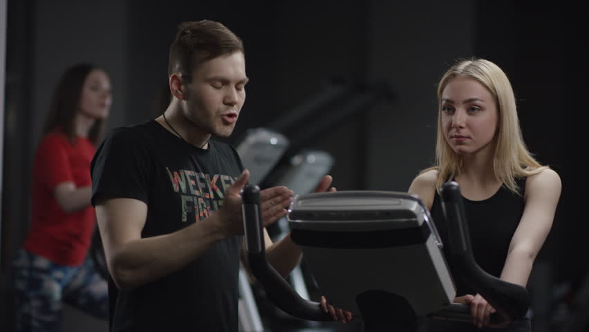 pretty girl in blak sport wear vigorously works on exercise bike and coach instructs about right way of exercise in the new gym against the background of engaged girls