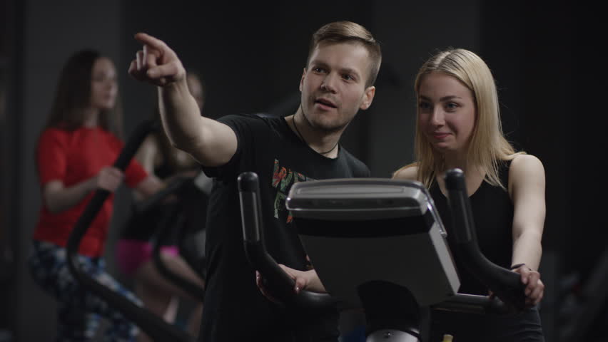 lovely girl in blak sport wear vigorously works on exercise bike and guy comes advise another trainer in the new gym against the background of engaged girls