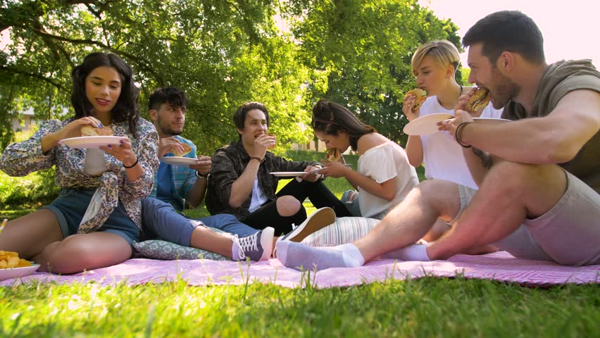 friendship, leisure and fast food concept - group of happy friends eating sandwiches or burgers at picnic in summer park