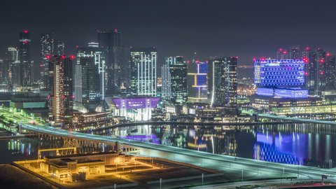 Aerial skyline of Abu Dhabi city centre from above during all night timelapse with illuminated skyscrapers and blinking light in windows. Light swithcing off. Traffic on the road.