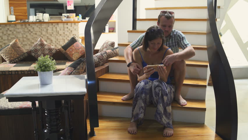 A man and a woman are sitting on the stairs in the house and watching movies on the smartphone Royalty-Free Stock Footage #1013232896