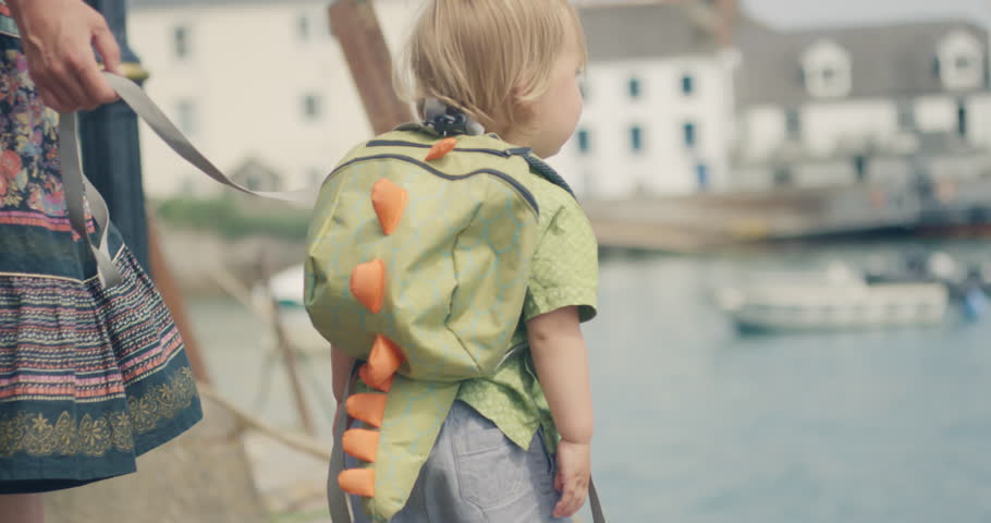 A little toddler boy in reins is walking with his mother by the water in a small village