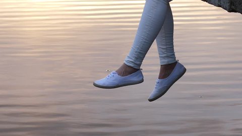 Swinging woman legs over quiet water surface. Enjoying loneliness concept. Woman dangling feet in white shoes over lake.