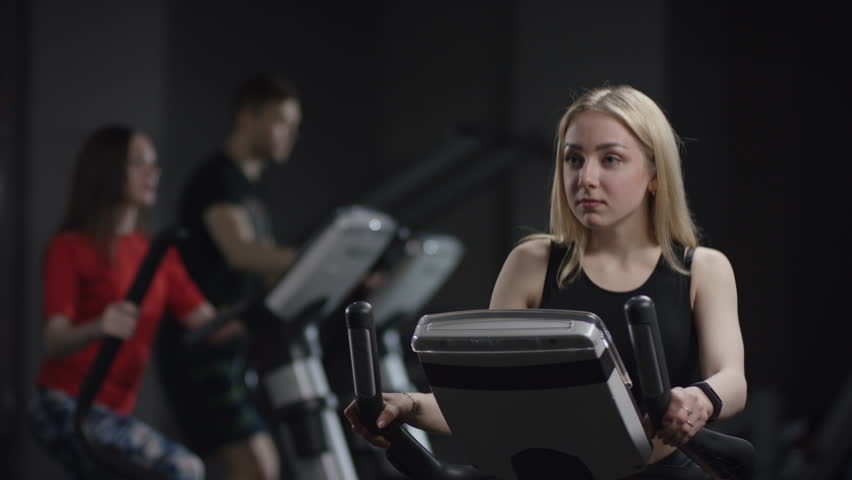 kind girl in blak sport wear vigorously works on exercise bike and watchs time with her wrist watch in the new gym against the background of engaged people