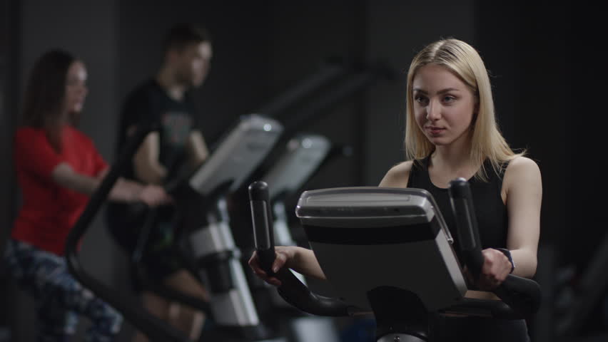 lovely girl in blak sport wear vigorously works on exercise bike and watchs time with her wrist watch in the new gym against the background of engaged people