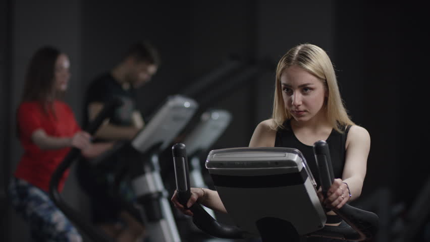 beautiful girl in blak sport wear vigorously works on exercise bike and watchs time with her wrist watch in the new gym against the background of engaged people