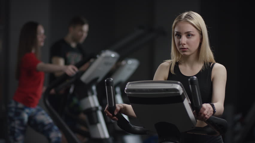pretty girl in blak sport wear vigorously works on exercise bike and watchs time with her wrist watch in the new gym against the background of engaged people