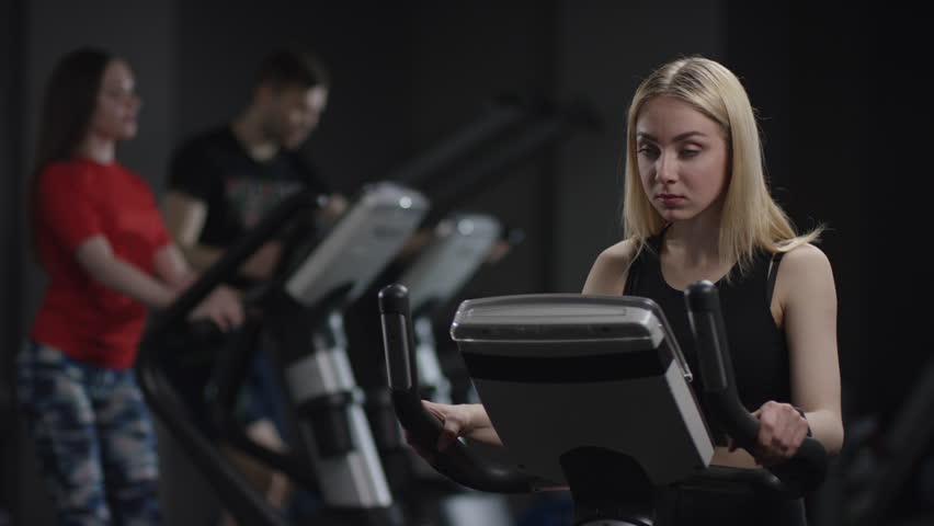 cute girl in blak sport wear vigorously works on exercise bike and watchs time with her wrist watch in the new gym against the background of engaged people
