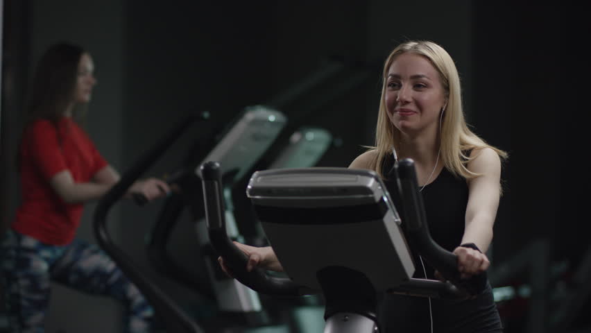 pretty girl in blak sport wear vigorously works on exercise bike and listens music with headphones and drink water in the new gym against the background of engaged people