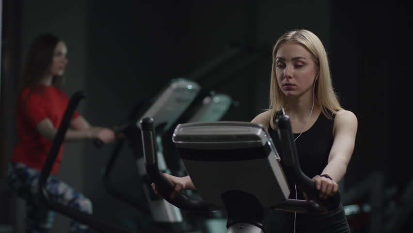honey girl in blak sport wear vigorously works on exercise bike and listens music with headphones and drink water in the new gym against the background of engaged people