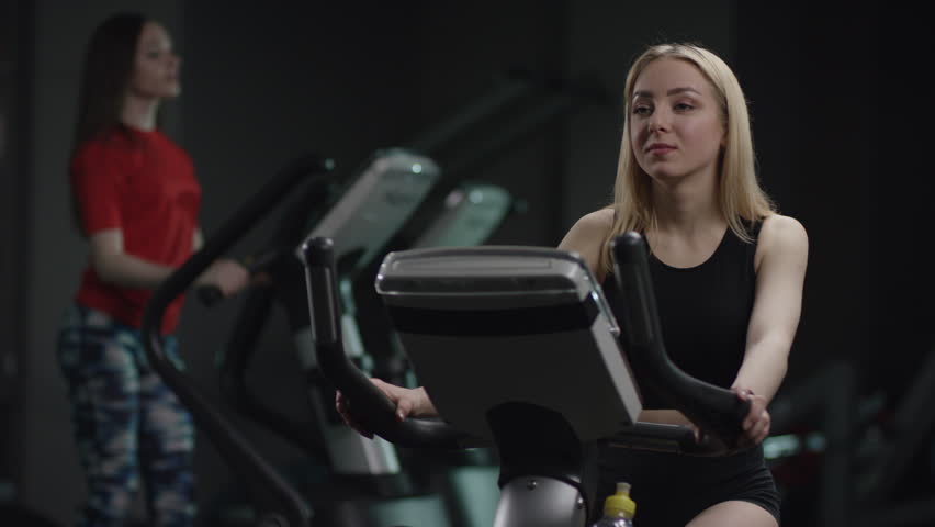 beautiful girl in blak sport wear vigorously works on exercise bike and listens music with headphones and drink water in the new gym against the background of engaged people