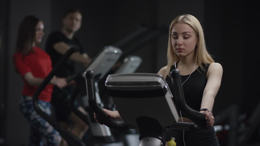 nice girl in blak sport wear vigorously works on exercise bike and listens music with headphones and drink water in the new gym against the background of engaged people