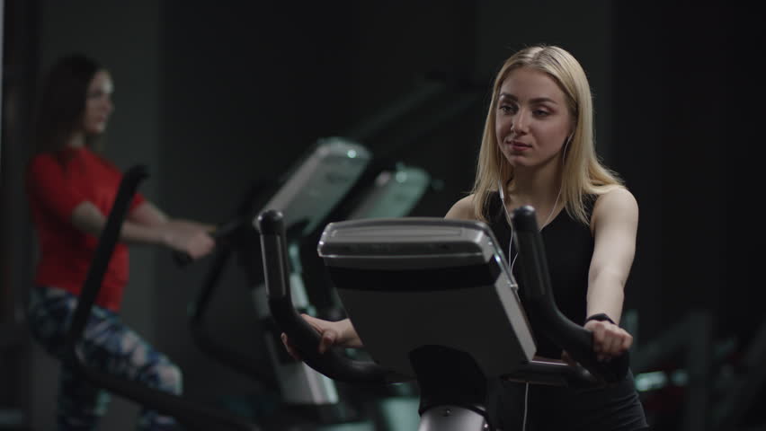 cool girl in blak sport wear vigorously works on exercise bike and listens music with headphones and drink water in the new gym against the background of engaged people