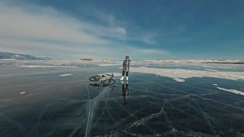 Woman is riding bicycle on the ice. The girl is dressed in a silvery down jacket, cycling backpack and helmet. Ice of the frozen Lake Baikal. The tires on the bicycle are covered with special spikes