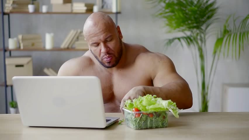Male bodybuilder sitting in the room at the laptop eating salad and answers...
