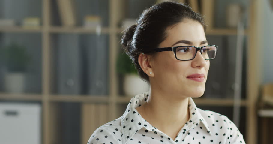 Close up of the Caucasian young beautiful woman in the white blouse and glasses working at the computer in the office and smiling. Portrait. Indoor