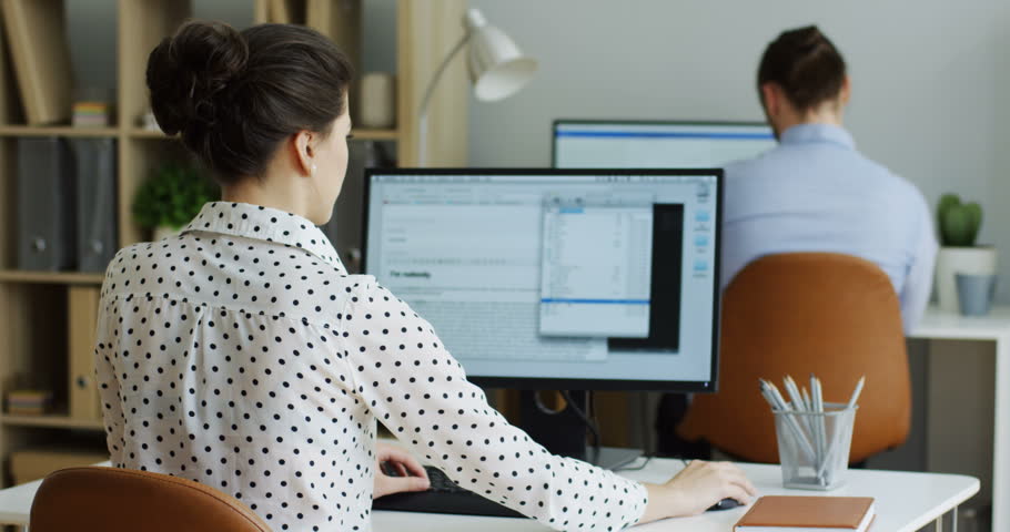Back view on the angry female office worker coping with a broken computer with an error writing on its screen, shaking it in nerves and male co-worker looking over shoulder. Indoor.