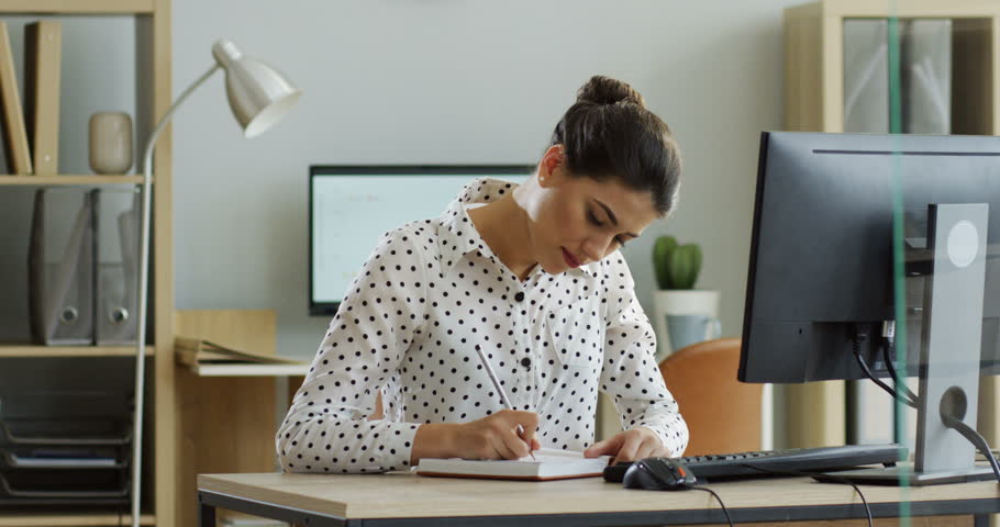 Young woman crossing out tasks in her notebook and preparing to go home, then her boss putting a lot of documents and work on her table. At office. Inside.