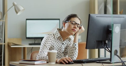 Young tired woman in glasses sitting at the laptop computer while working in the office, then almost falling asleep and waking up. Indoor.