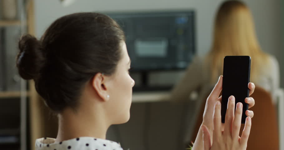 Rear of the brunette young woman sitting at her workplace in the office and using her smartphone with broken screen, cannot do anything with device and being nervous. Indoors.