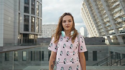 Slow Motion Portrait of Attractive Cute Smiling Caucasian Ethnicity Young Woman with Headphones and Backpack in Urban Environment. Millenial Hipster New Generation Y.