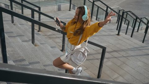 Portrait of young cute attractive young girl in urban city streets background listening to music with headphones. Woman wearing yellow blouse and silver skirt.
