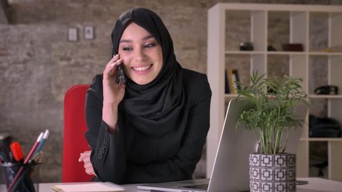 Young beautiful muslim girl in hijab is talking on smartphone in office, work concept